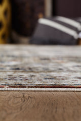 Close-up of the Aged Treasures Premium Rug - 2181A, a light geometric design on wood flooring. The image highlights the machine-made rug's edge and floor texture, with blurred striped pillows and dark furniture in the background.
