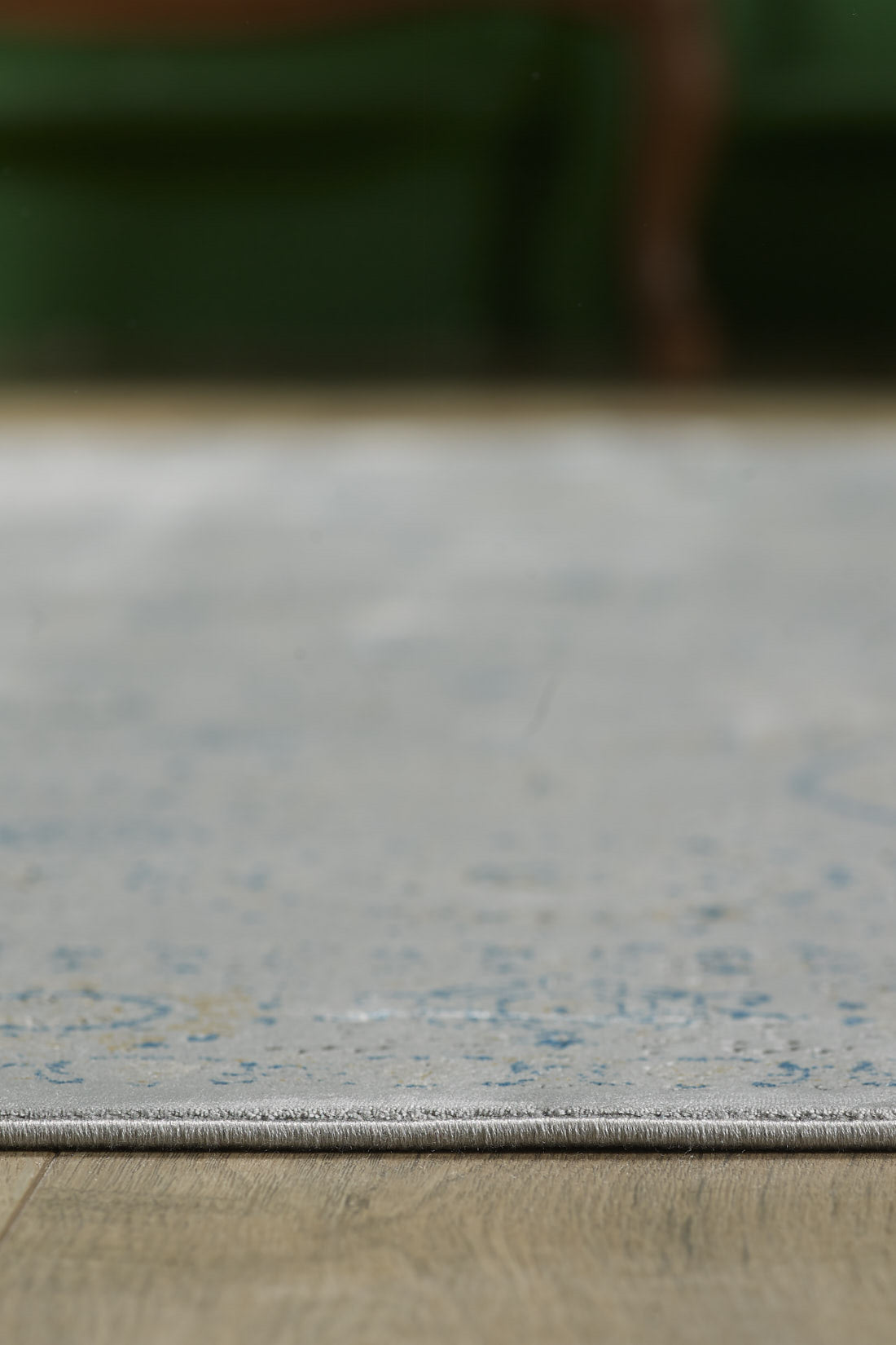 A close-up of the Ancestral Motifs Turkish Rug - 2225C, showcasing blue patterns on modal silk-polyester, placed on a wooden floor with a blurred backdrop featuring green and brown furniture.