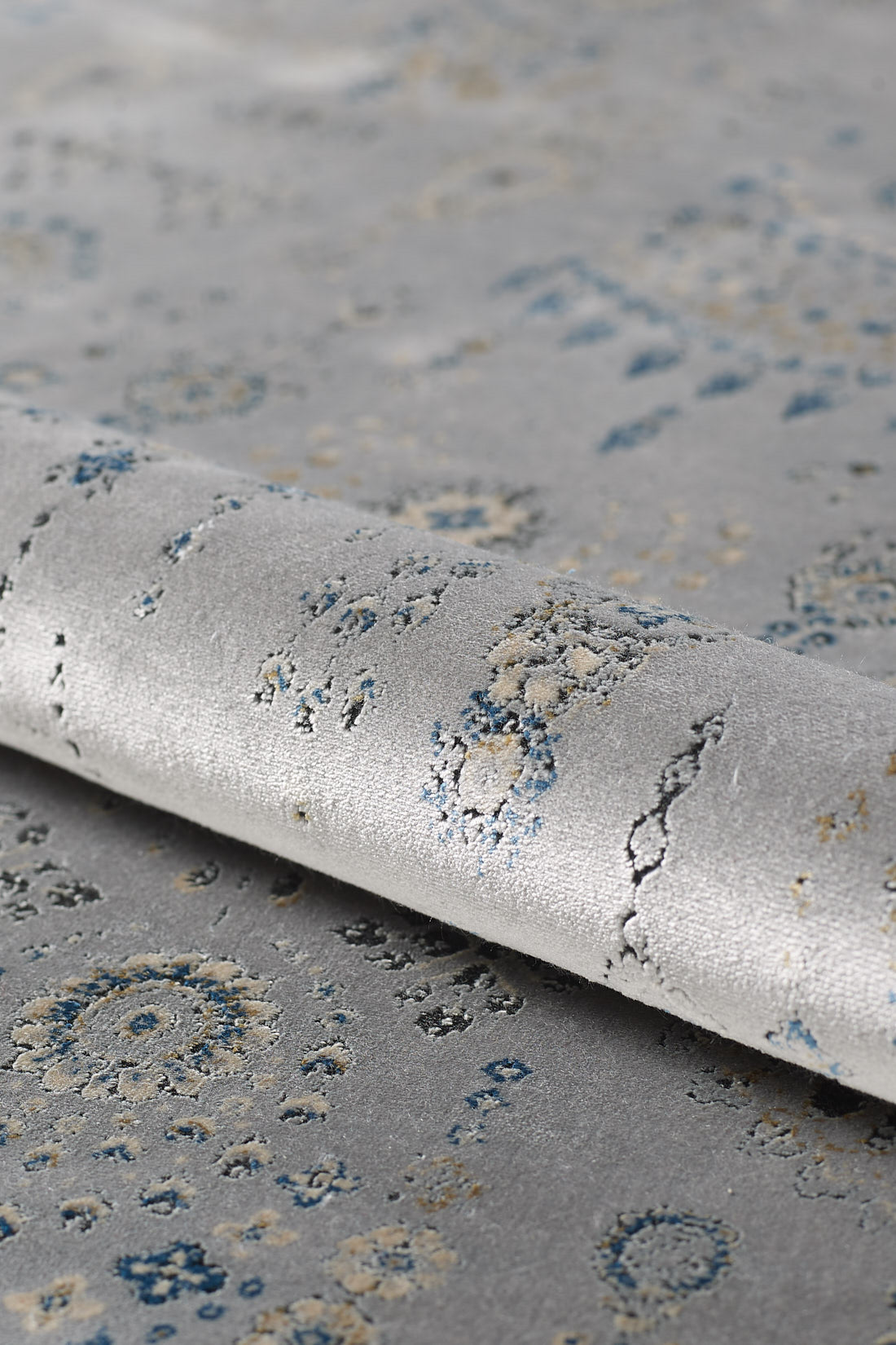 A close-up of the Ancestral Motifs Turkish Rug - 2225C in light gray, featuring blue and beige abstract patterns, crafted from modal silk-polyester with a subtly rolled edge and textured surface.