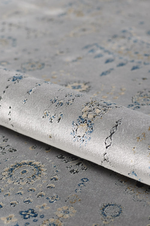 Load image 1 in gallery view. A close-up of the Ancestral Motifs Turkish Rug - 2225C in light gray, featuring blue and beige abstract patterns, crafted from modal silk-polyester with a subtly rolled edge and textured surface.