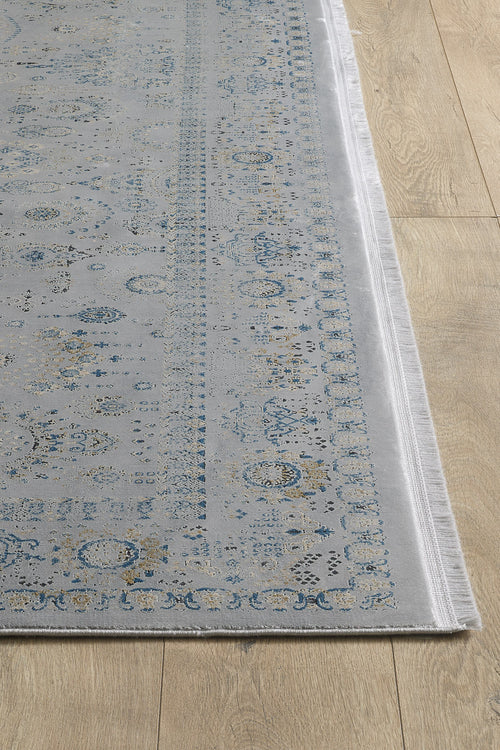 Load image 1 in gallery view. A close-up of the Ancestral Motifs Turkish Rug - 2225C, a contemporary machine-made rug in light gray with blue and beige floral patterns, crafted from modal silk-polyester, bordered decoratively with fringed edges on a light wood floor.