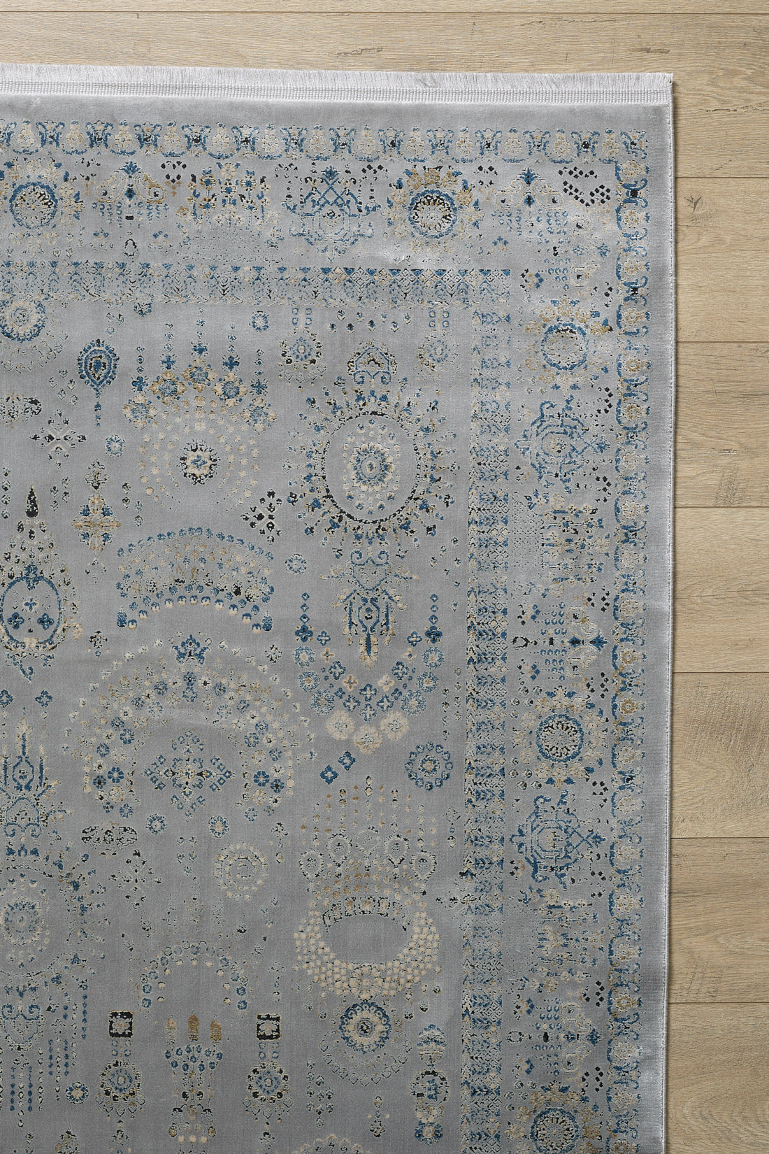A corner of the Ancestral Motifs Turkish Rug - 2225C, a modern machine-made rug in blue, cream, and gold on light gray, lies on a light wood floor. Crafted from modal silk-polyester with a decorative border and worn-effect detailing.
