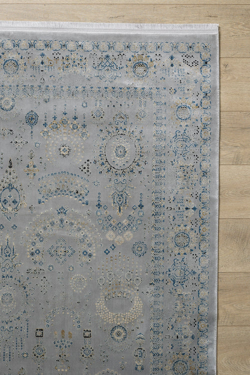 Load image 1 in gallery view. A corner of the Ancestral Motifs Turkish Rug - 2225C, a modern machine-made rug in blue, cream, and gold on light gray, lies on a light wood floor. Crafted from modal silk-polyester with a decorative border and worn-effect detailing.