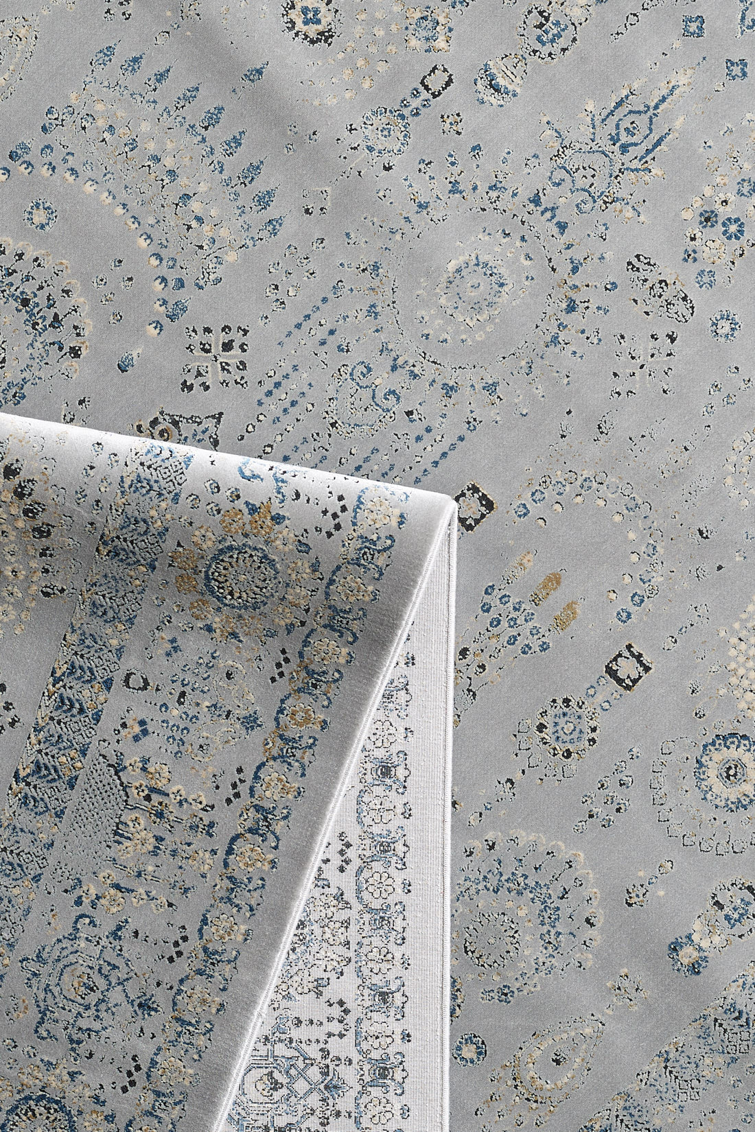 The Ancestral Motifs Turkish Rug - 2225C is a machine-made rug with blue, beige, and gray geometric and floral patterns. A folded corner shows the lighter underside, echoing the rug's intricate contemporary design.