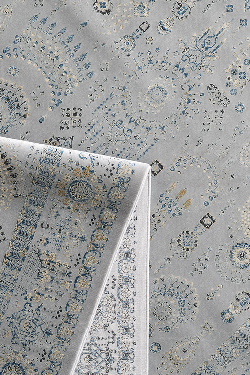 Load image 1 in gallery view. The Ancestral Motifs Turkish Rug - 2225C is a machine-made rug with blue, beige, and gray geometric and floral patterns. A folded corner shows the lighter underside, echoing the rug's intricate contemporary design.