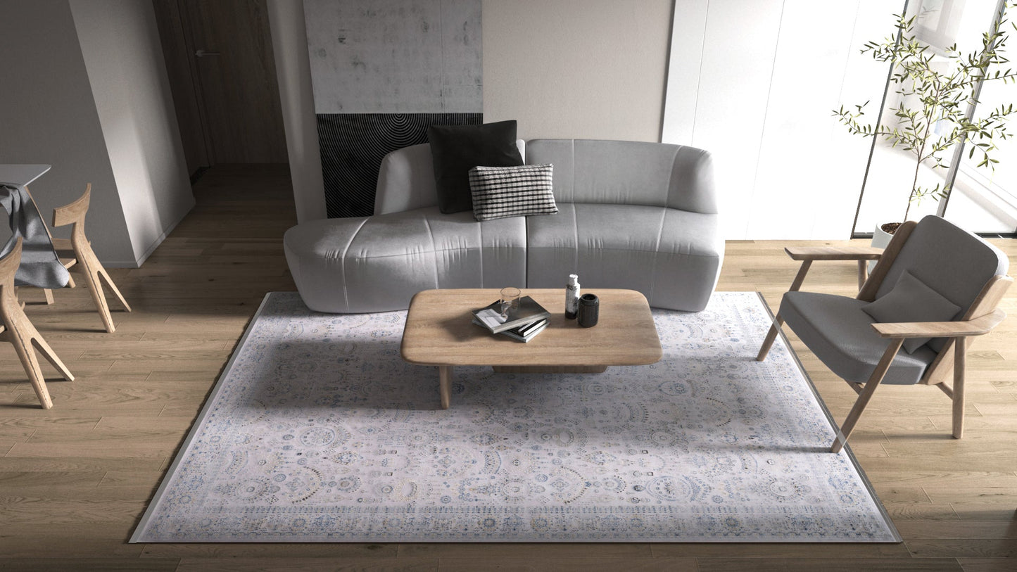 A modern living room with a gray sofa, wooden coffee table, armchair, and the Ancestral Motifs Turkish Rug - 2225C. Sunlight highlights the wooden flooring and a tall potted plant in the minimalist decor.