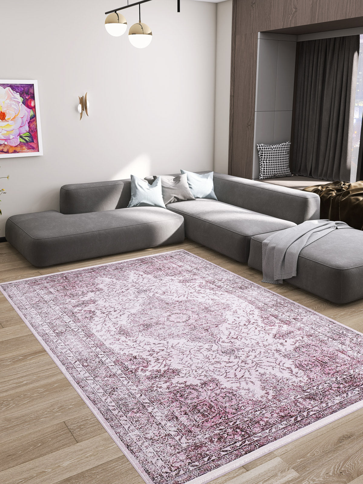 A modern living room features a large grey sectional, light grey and blue pillows, the Ancient Emblem - Washable Rug - JR200 (Custom Size) in pink on wooden flooring, wall art, a blanket, and a window nook with dark curtains.