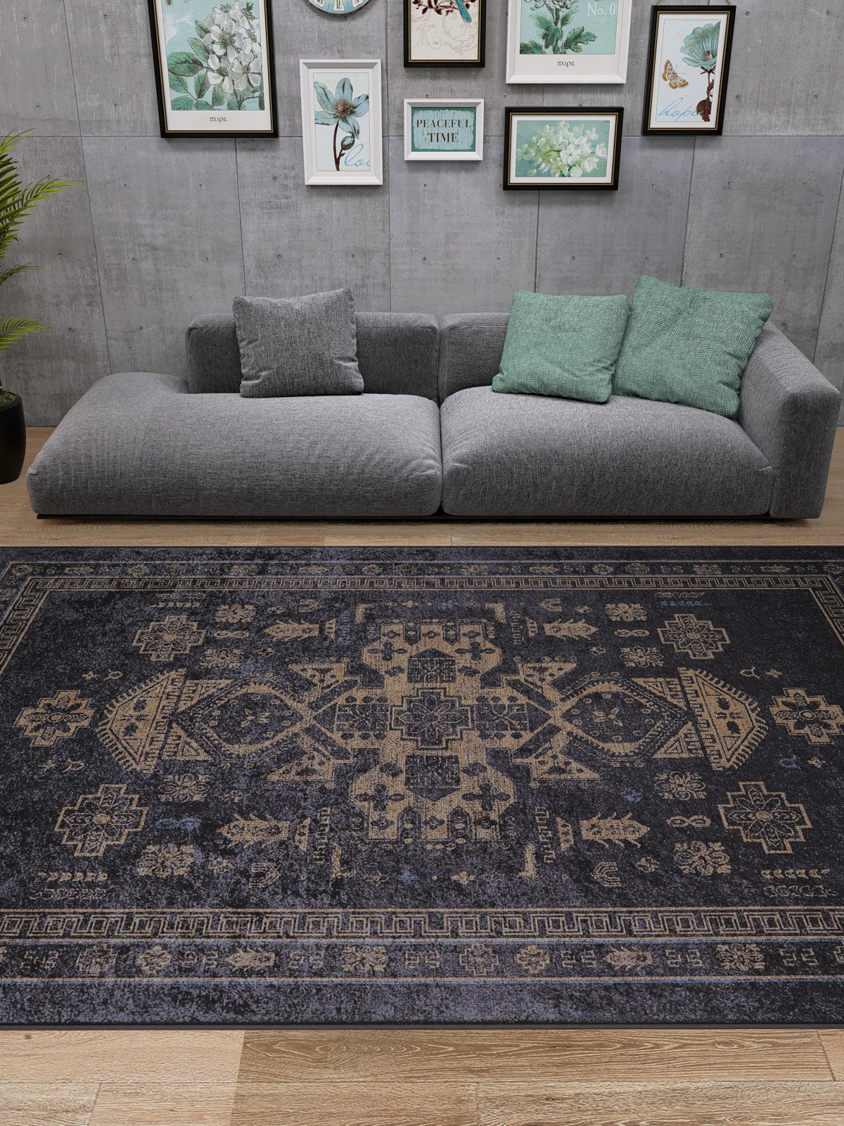 A modern living room features a gray sectional, green and gray pillows, framed botanical art on a concrete wall, and the Ancient Legacy - Washable Rug - JR1890 (Custom Size) in black. A potted plant sits to the left on wooden flooring.