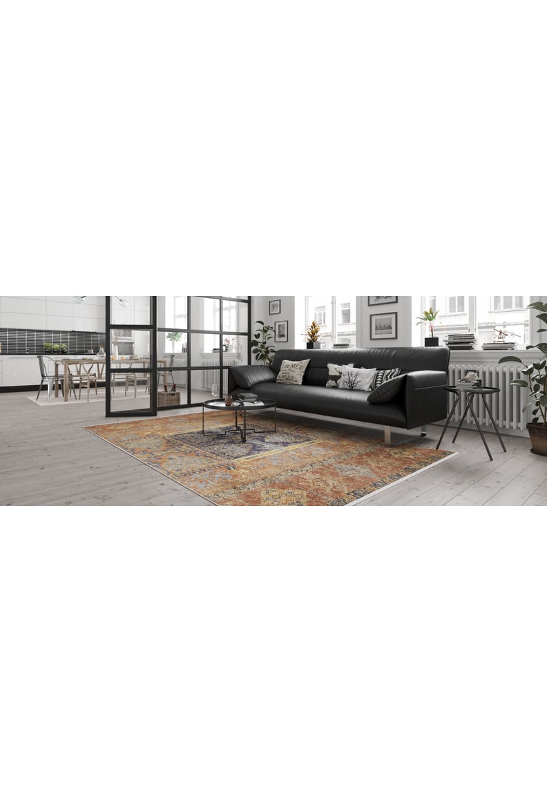 A modern living room features a black sofa, patterned pillows, and the Ancient Palace - Washable Rug - JR1864 (Custom Size) in orange—ideal for high-traffic areas. A small coffee table, large windows, plants, plus a dining area and white-cabinet kitchen complete the space.