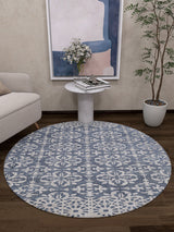 The Antique Flourish - Washable Rug - JR1591, in round blue and white, sits on a dark wood floor beside a light armchair. A small white side table holds books and flowers, with a potted plant and abstract wall art—perfect for high-traffic areas.