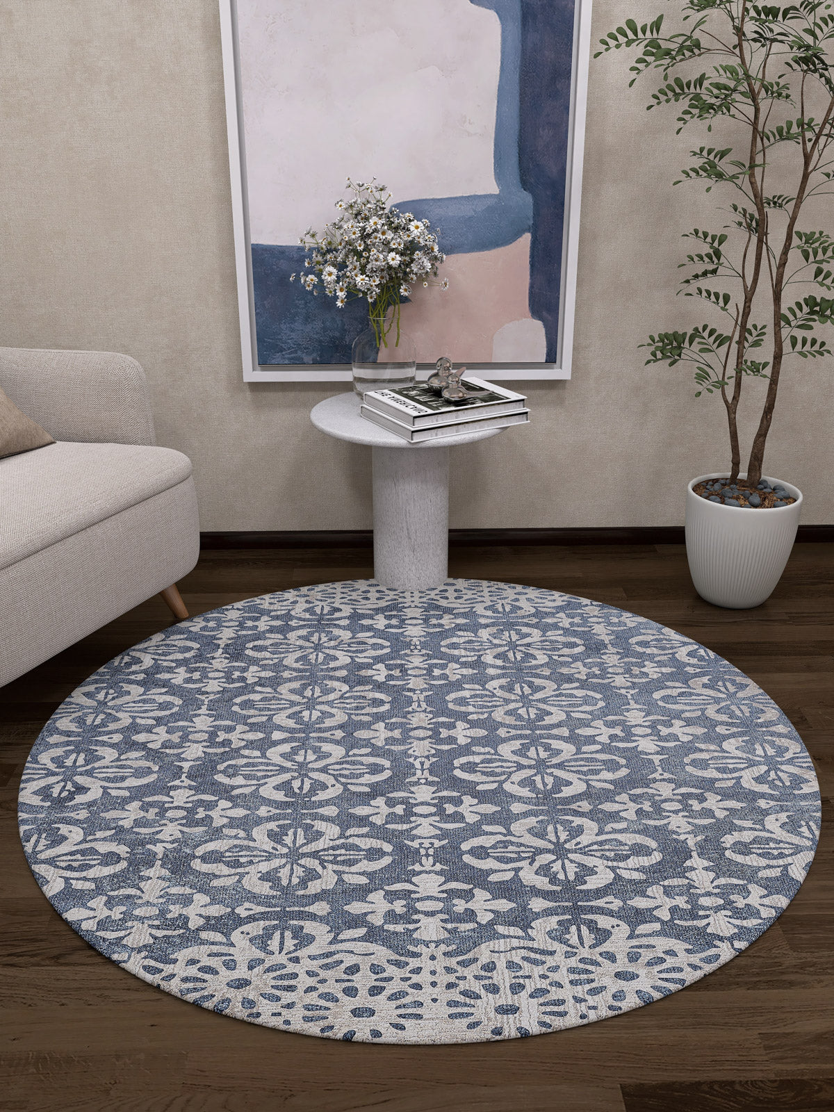 The Antique Flourish - Washable Rug - JR1591 (Custom Size) features a round blue and white pattern, ideal for cozy living rooms or high-traffic areas, shown on a dark wooden floor beside a light sofa, side table with books, plant, and abstract wall art.