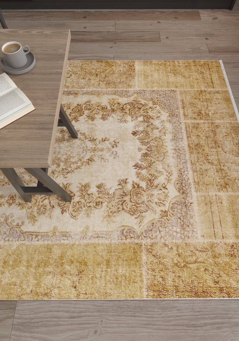 The Antique Insignia - Washable Rug - JR309 (Custom Size), featuring beige and yellow-gold floral patterns, lies on a wooden floor by a table with coffee and a book—ideal for high-traffic areas.