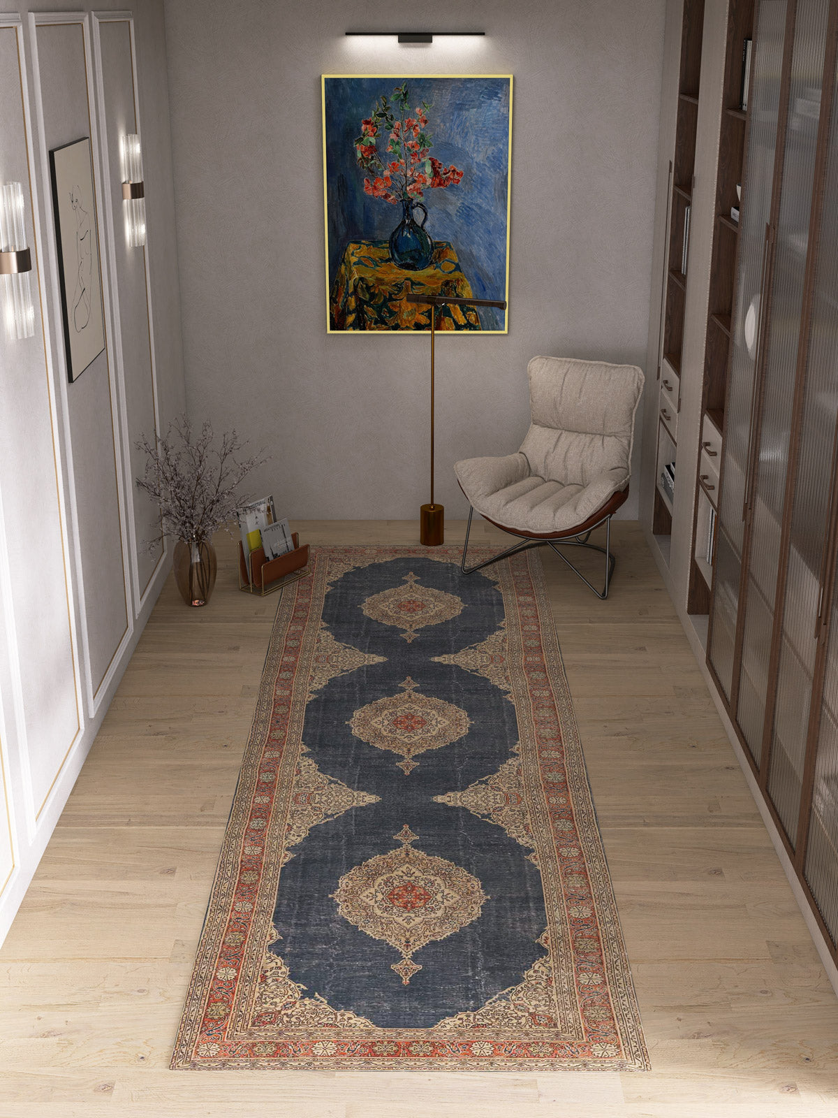 A modern hallway with light wood floors features the Antique Symphony - Washable Rug - JRN1017 (Custom Size) with a medallion design, paired with a cushioned chair, glass vase of dried branches, wall shelves, and a framed floral painting under a spotlight.