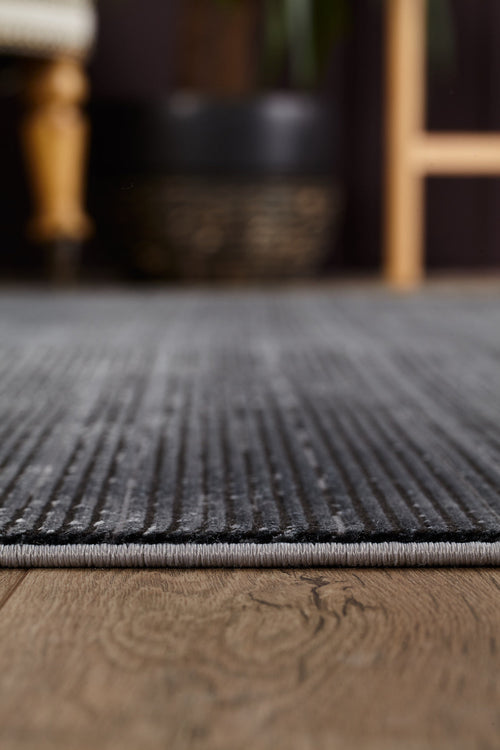 Load image 1 in gallery view. Close-up of the Antrachite Shadows Black Rug - M497A, a grey machine-made rug with subtle stripes, shown on a wooden floor. Blurred background reveals parts of furniture, including a chair leg and a plant pot.