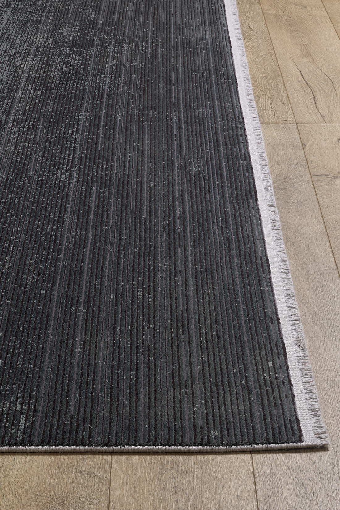 A close-up of the Antrachite Shadows Black Rug - M497A, a dark gray, ribbed machine-made rug with light fringe edges on a light wood floor. The minimalist design features subtle vertical textures for a modern look.