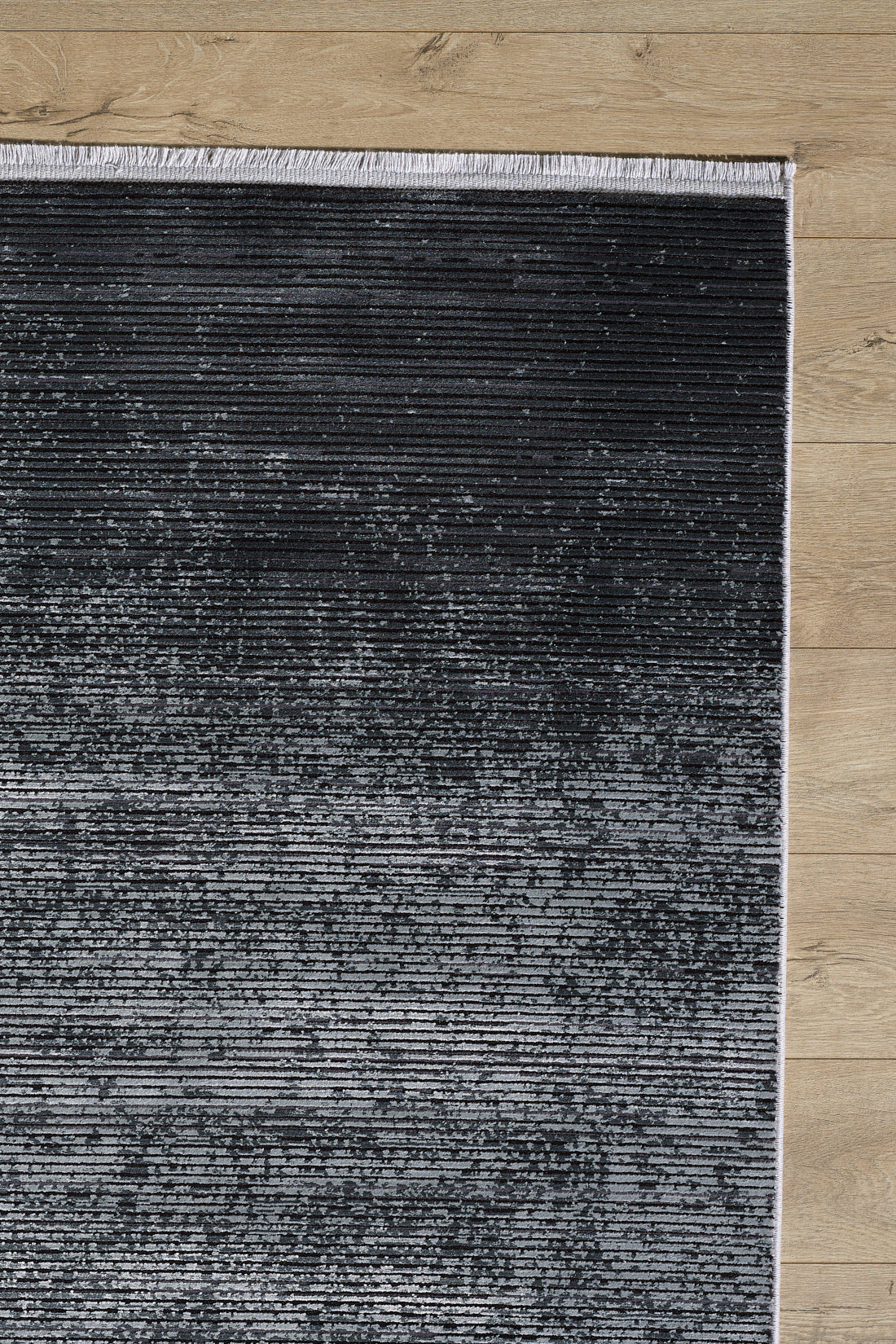 The Antrachite Shadows Black Rug - M497A features a modern gradient from dark to light gray with subtle stripes, visible fringe on the top edge, and a minimalist machine-made design—shown on a light wooden floor.