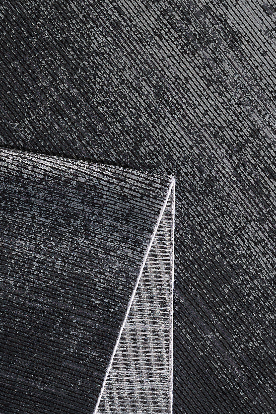 Close-up of a textured black and silver surface with diagonal lines. The Antrachite Shadows Black Rug - M497A, featuring a matching linear pattern, sits in the bottom left corner, creating contrast against the background.