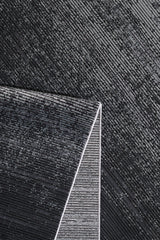 Close-up of a textured black and silver surface with diagonal lines. The Antrachite Shadows Black Rug - M497A, featuring a matching linear pattern, sits in the bottom left corner, creating contrast against the background.
