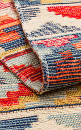 A close-up of the Aqua Flow - Kilim - NMD02 reveals blue tones with red, orange, and beige geometric patterns, showcasing the allergy-friendly rug’s textured stitching along its folded edge.