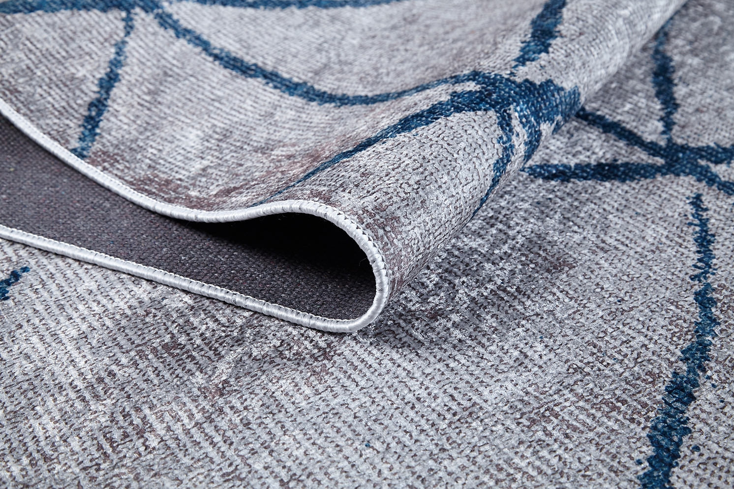 A close-up of the Arabesque Trellis - Washable Rug - JR1732 (Custom Size) corner folded over, featuring a textured grey surface with blue geometric lines and dark underside—ideal for high-traffic areas.