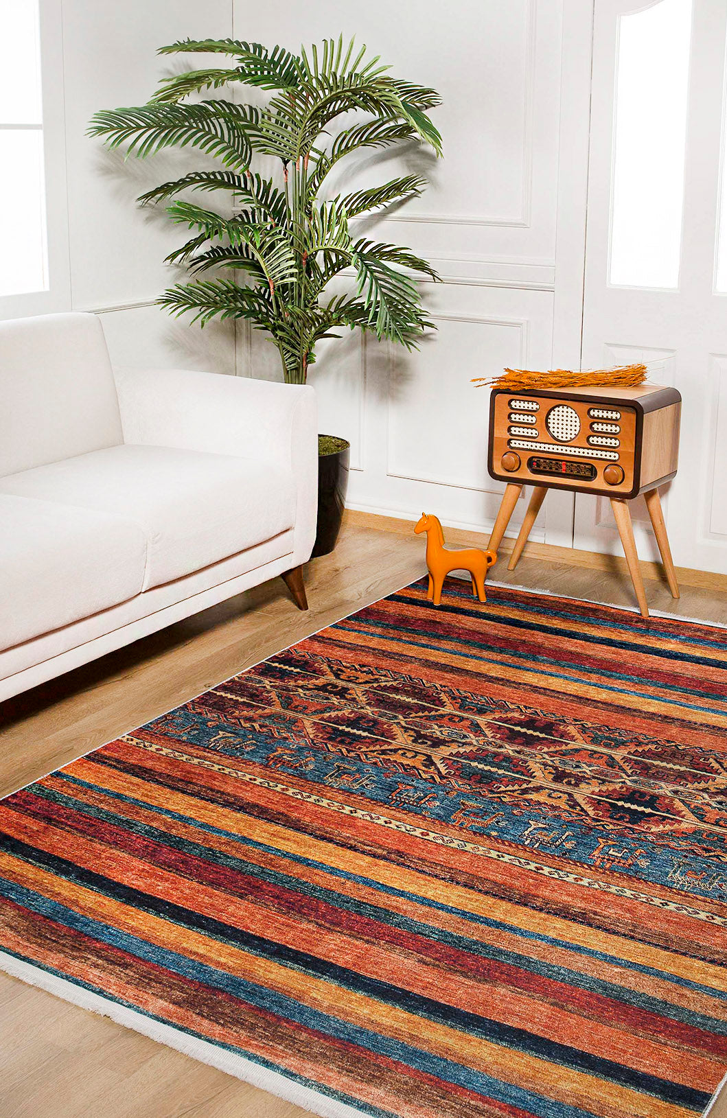 A cozy living room features the Arctic Chill - Washable Rug - JR860 (Custom Size), ideal for high-traffic areas, along with a white sofa, potted plant, orange decorative horse, vintage radio on a wooden stand, and bright natural light.