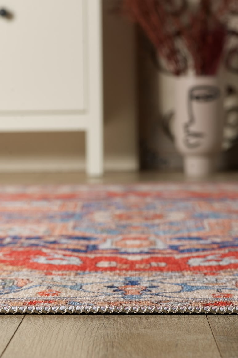 Close-up of the Ardentis Medallion Washable Rug - LCC3002 (Outlet) US in red and multicolor on a wooden floor, with a white cabinet and face-design vase blurred in the background. This washable rug adds artful style to your space.