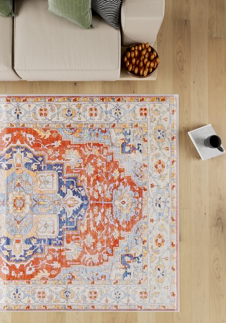 A top-down view of the Ardentis Medallion Washable Rug - LCC3002 (Custom Size) in red multicolor with blue and beige designs, placed next to a beige sofa, a bowl of dates, and a mug on an open book on a light wooden floor.