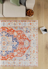 A top-down view of the Ardentis Medallion Washable Rug - LCC3002 (Custom Size) in red multicolor with blue and beige designs, placed next to a beige sofa, a bowl of dates, and a mug on an open book on a light wooden floor.