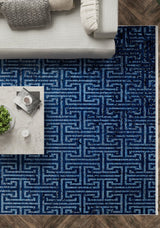 A top-down view of a living room featuring the Art Deco Elegance - Washable Rug - JR1224 with a blue geometric pattern—perfect for high-traffic areas—a marble coffee table with books and a plant, plus a light sofa adorned with patterned and plain pillows.