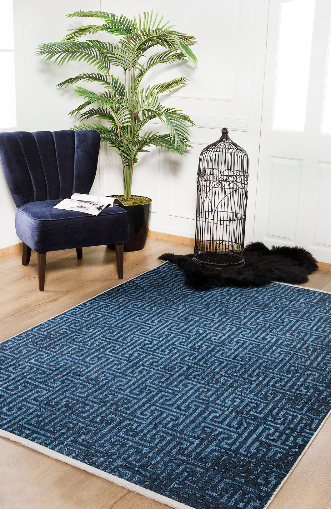A modern room features the Art Deco Elegance - Washable Rug - JR1224 (Custom Size), a navy velvet chair with a book and glasses, a potted plant, black faux fur rug, and empty birdcage—ideal for high-traffic areas.