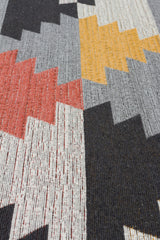A close-up of the Artisan Touch - Washable Double-Sided Kilims - AR20 rug shows a geometric zigzag pattern in black, gray, yellow, red, and white. The recycled cotton weave highlights the interlaced threads and textured fabric.