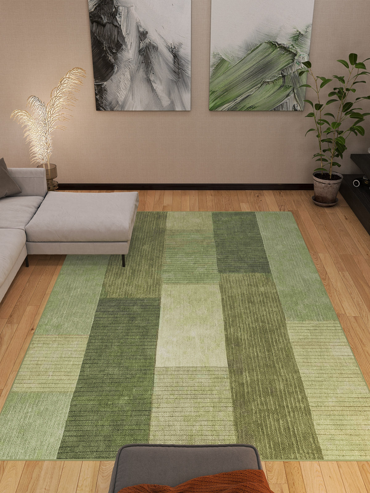 A modern living room features the Artisanal Essence - Washable Rug - JRN32 (Custom Size) with a green checkered pattern on a wooden floor, paired with a gray sectional sofa, abstract wall art, a potted plant, and a feather lamp in the corner.