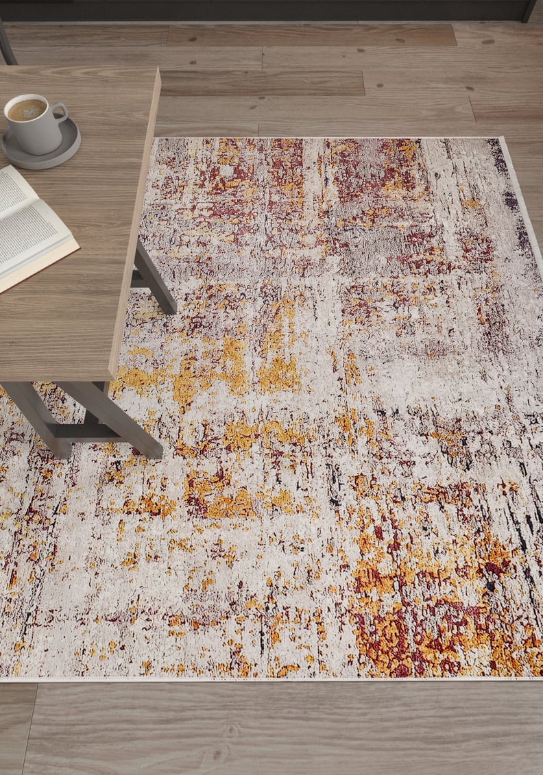 The Artistic Abstraction - Washable Rug - JR775 (Custom Size), featuring a modern abstract design in white, yellow, and burgundy, rests on a wood floor beside a light wood table with an open book and coffee cup.