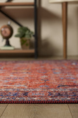 A close-up of the Aurelius Coral Washable Rug - LCC3005, featuring red and purple patterns on a wooden floor, perfect for high-traffic areas, with blurred shelves, a plant, and decor in the background.