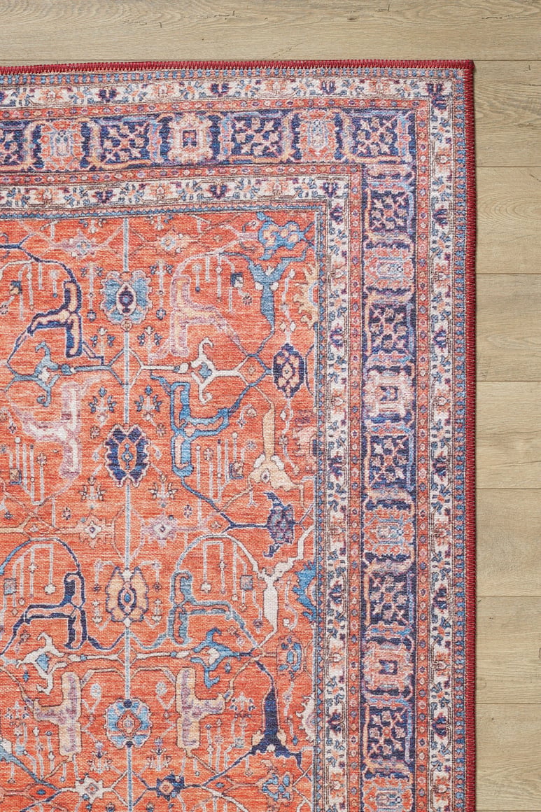 A corner of the Aurelius Coral Washable Rug - LCC3005, featuring vibrant orange, blue, and cream geometric and floral patterns, lies on a light wood floor. This durable flat weave style is ideal for high-traffic areas.