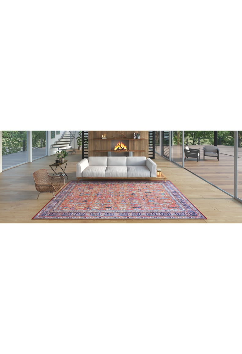 A modern living room with large windows, a white sofa, wooden accent wall and fireplace, a washable Aurelius Coral Rug - LCC3005 (Custom Size) in pink and purple, a wicker chair, and greenery visible outside.