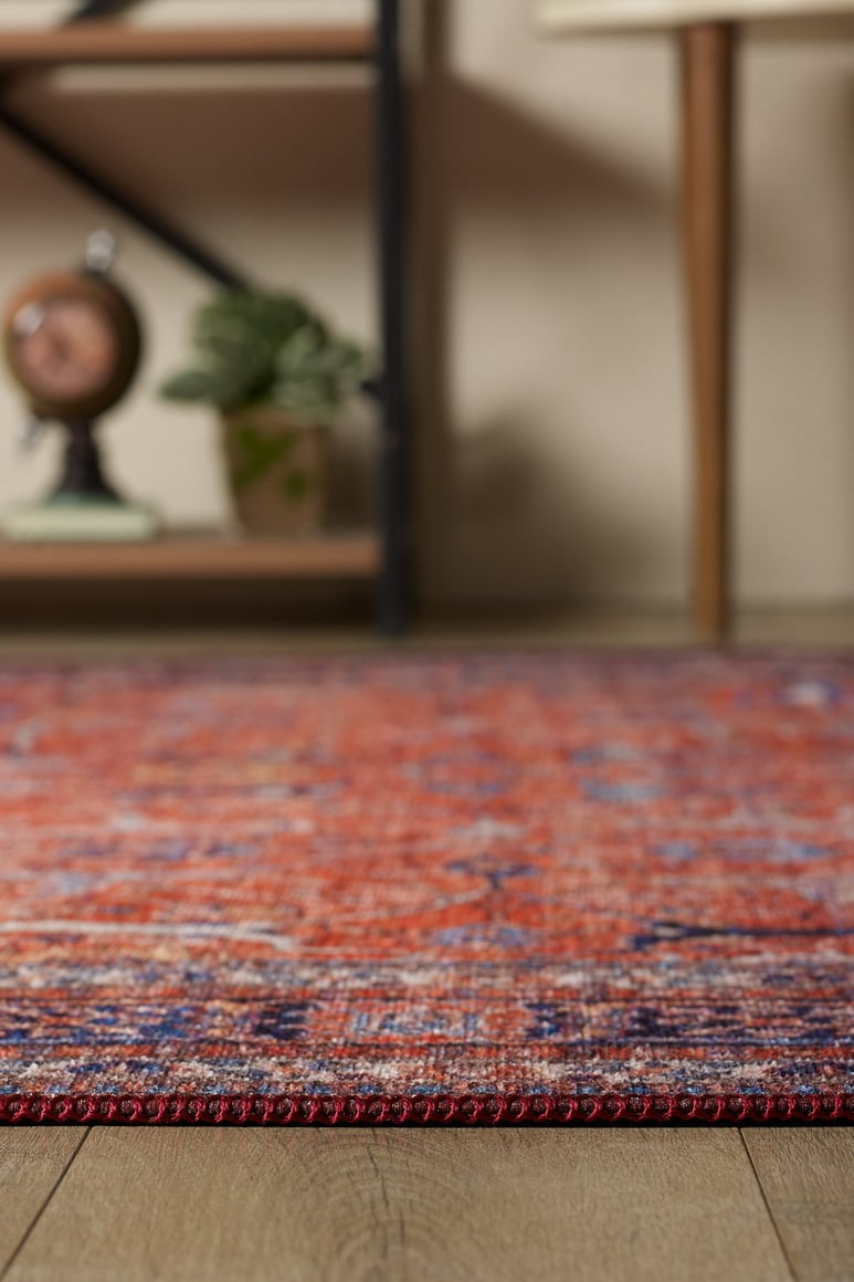 A close-up of the Aurelius Coral Washable Rug - LCC3005 (Custom Size) with a red and purple pattern on a wooden floor, blurred shelves and furniture in the background—perfect for allergy-sensitive or high-traffic areas.