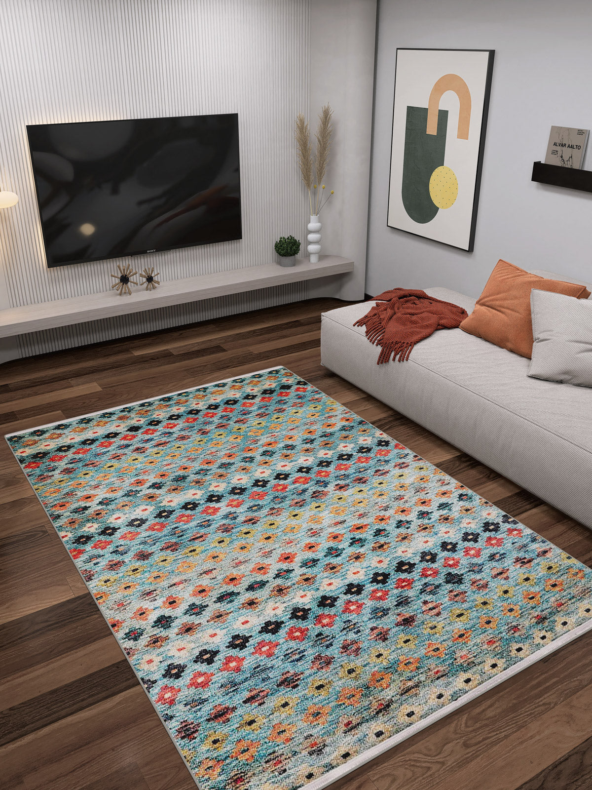 A modern living room featuring the Aztec Ambience - Washable Rug - JR1086, a low gray sectional with pillows and an orange throw, wall art, flat-screen TV, wood flooring, and minimalist decor—ideal for high-traffic spaces.