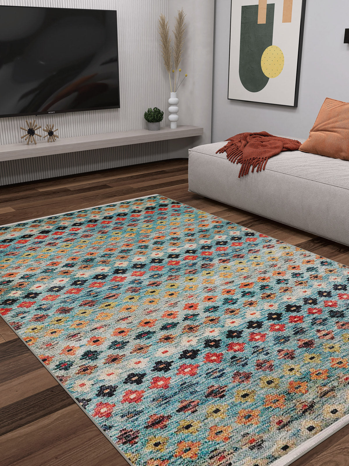 A modern living room featuring the Aztec Ambience - Washable Rug - JR1086 (Custom Size), ideal for high-traffic areas, a low beige sofa with an orange throw, wall-mounted TV, minimalist art, pampas grass vases, and wood flooring.