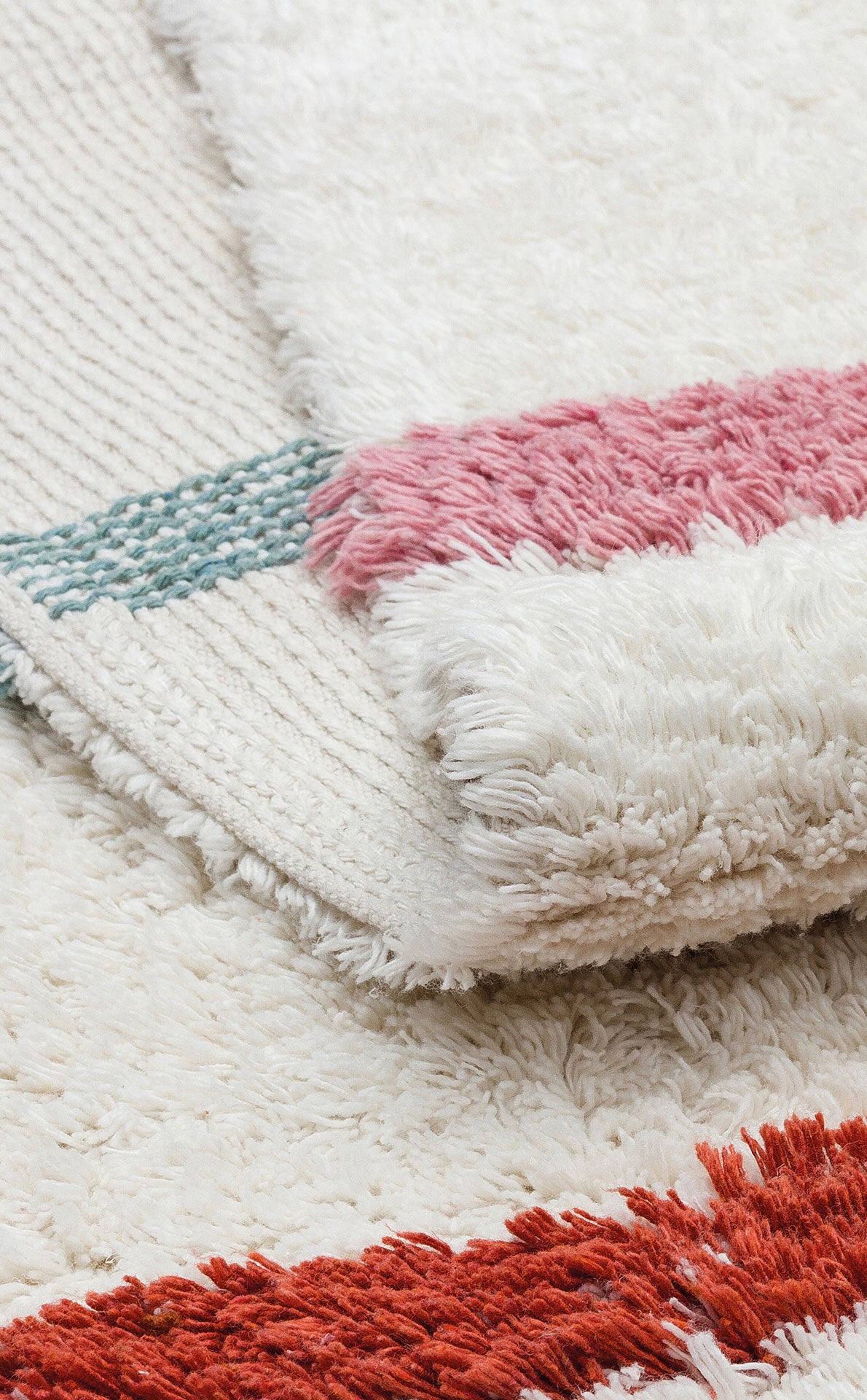 A close-up of the Baby Breeze - Washable Shaggy - CBN01 rug with a folded corner, featuring woven blue and pink stripes and thick red shaggy fibers—perfect as a cozy kids room rug.