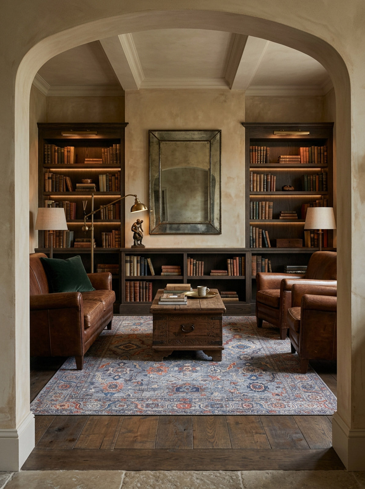 A cozy library with leather armchairs, a wooden coffee table, bookshelves, table lamps, and a large mirror features the Bellaris Gray Washable Rug - LCC3003 for easy care, all viewed through an arched doorway.