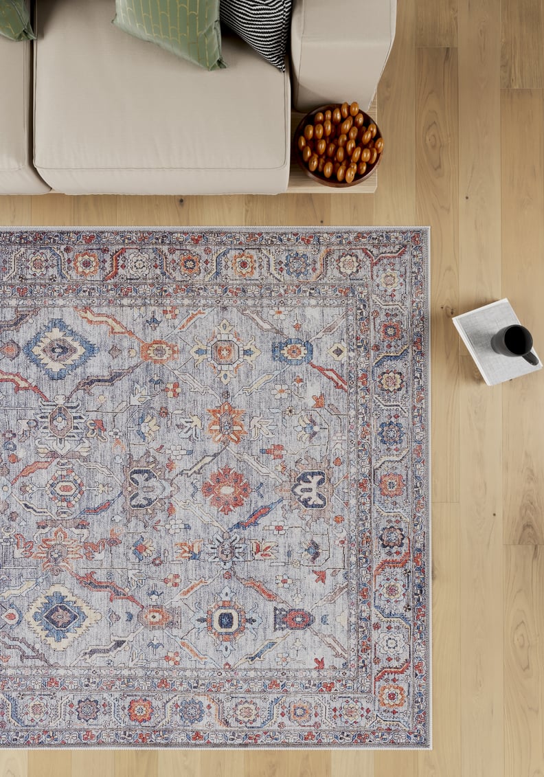 The Bellaris Gray Washable Rug - LCC3003 (Outlet) US, featuring blue, orange, and beige designs, lies on a wooden floor beside a beige sofa with green and black pillows. Nearby are a bowl of breadsticks and an open magazine with a cup.