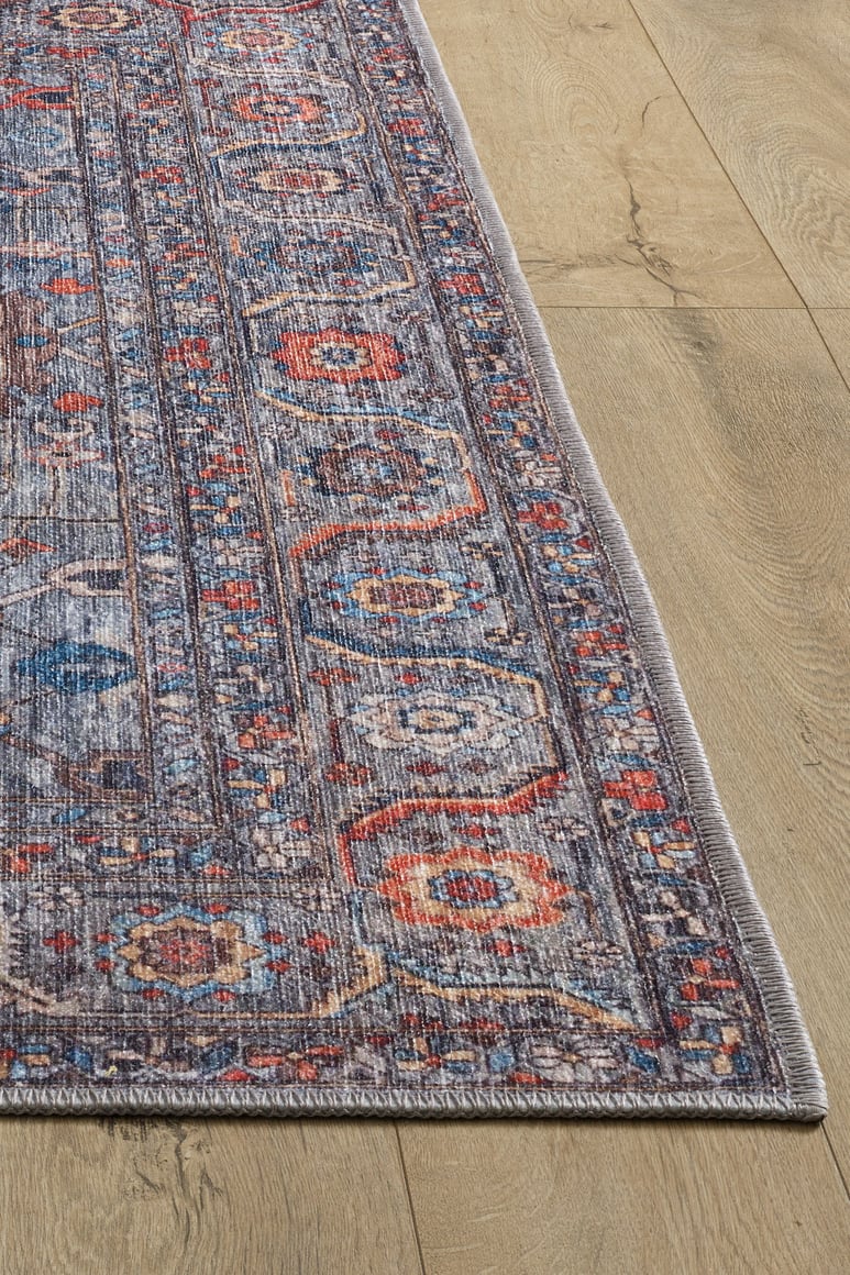 A close-up of the Bellaris Gray Washable Rug - LCC3003 (Outlet) US, featuring intricate geometric and floral patterns in blue, red, and beige on a light wood floor. The rug’s woven texture is visible along the edge.