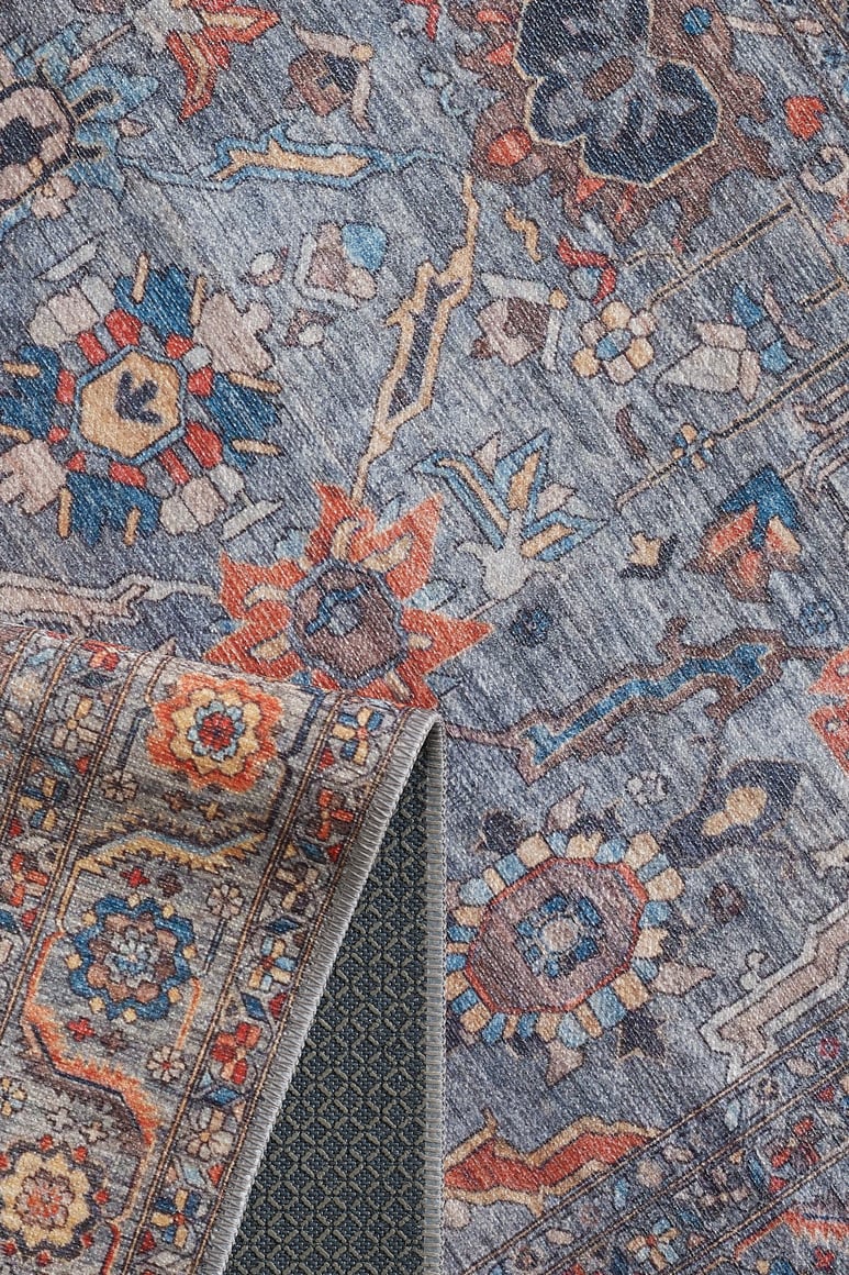 A close-up of the Bellaris Gray Washable Rug - LCC3003 (Outlet) US shows a blue-gray background with intricate floral and geometric patterns, plus a folded corner revealing its textured dark blue and gray lattice underside.
