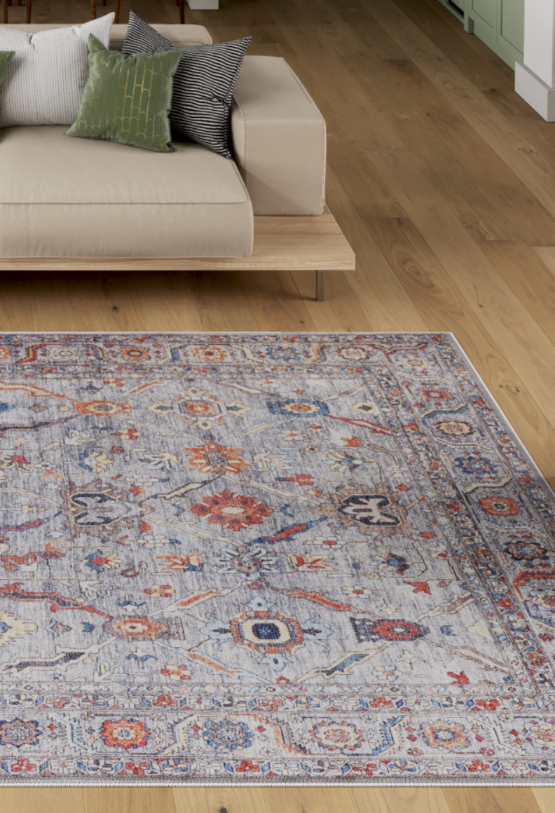 A modern living room with a light wood floor, a beige minimalist sofa with three green and black cushions, and a large Bellaris Gray Washable Rug - LCC3003 (Custom Size) in blue, red, and beige tones.