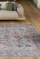 A modern living room with a light wood floor, a beige minimalist sofa with three green and black cushions, and a large Bellaris Gray Washable Rug - LCC3003 (Custom Size) in blue, red, and beige tones.