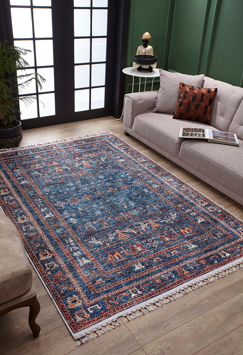 A living room with the Berber Beauty - Washable Rug - JR5092 (Custom Size), blue with red and cream patterns on wooden floors, a light gray sofa, round side table, green walls, and large windows—ideal for high-traffic spaces.
