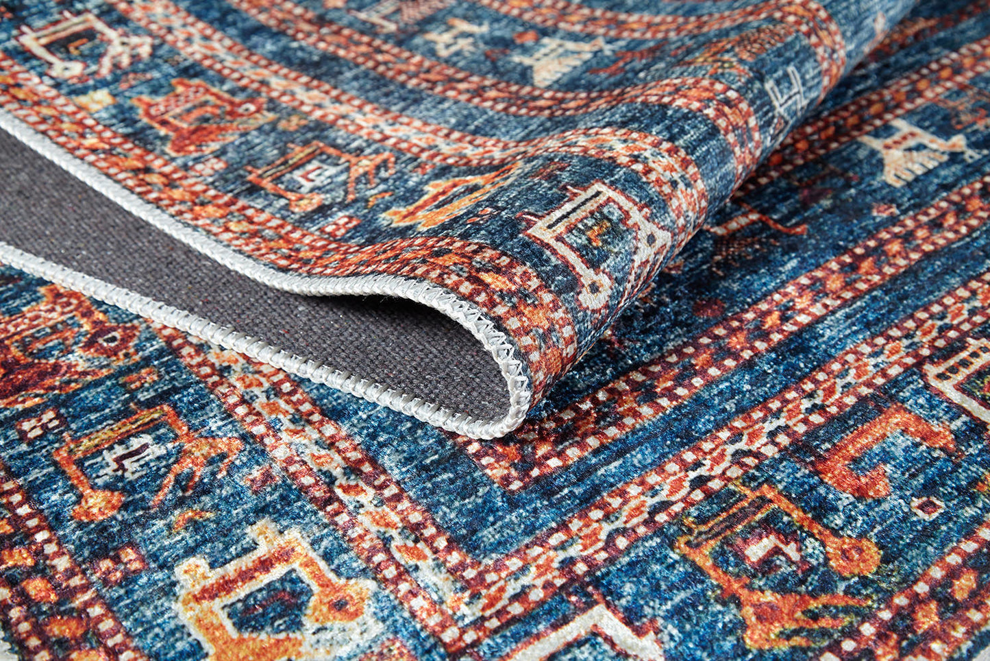 A close-up of the Berber Beauty - Washable Rug - JR5092 (Custom Size) showcases its blue, red, and beige geometric patterns. The folded edge reveals a stitched border—durable and ideal for high-traffic areas.