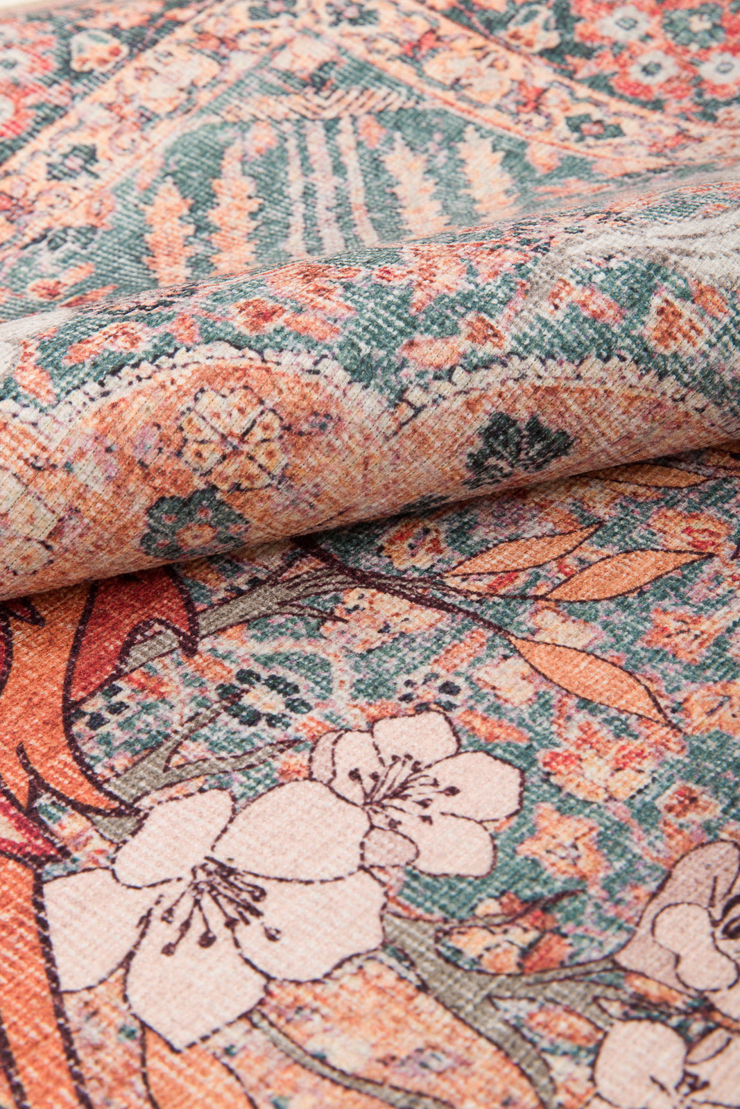 A close-up of the Blazefang Myth - Washable Rug (JR0049), a flat weave rug with intricate floral and geometric designs in muted orange, green, cream, and brown tones, shown with one edge slightly folded.