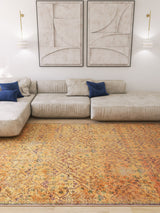 A modern living room with a beige sectional sofa, blue accent pillows, abstract neutral wall art, wall-mounted sconces, and the Blossom Breeze - Washable Rug - JR1752 in orange covering the floor.